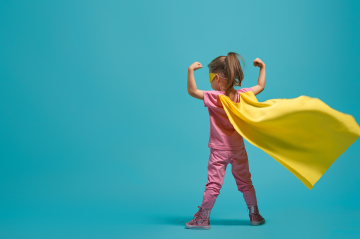 Ein kleines Mädchen steht vor einem blauen Hintergrund mit gelben Umhang in Superman - Manier.