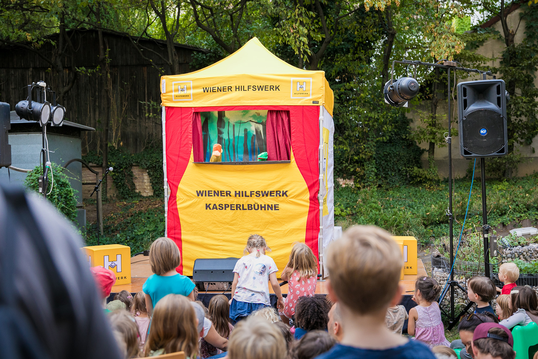 Kinder sitzen im Wiener Hilfswerk Garten und schauen einer Kasperlvorstellung zu