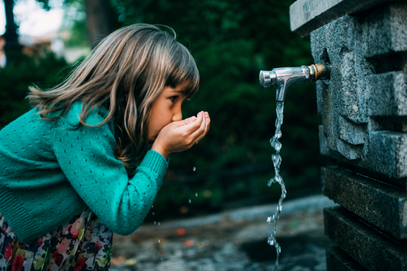 Ein Mädchen trinkt aus einem Brunnen kühles Wasser.