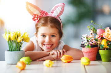 ein lachendes Mädchen trägt einen Haarreifen mit bunten Hasenohren und hat die Arme auf einem Tisch abgelegt. Auf dem Tisch sind Töpfe mit Frühlingsblumen und Ostereier 
