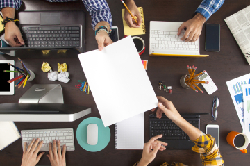 Ein Schreibtisch mit Computern und Dokumenten und Menschen die Arbeiten 