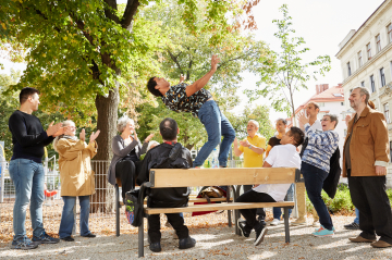 Eine Gruppe beklatscht einen Mann der auf einer Parkbank tanzt