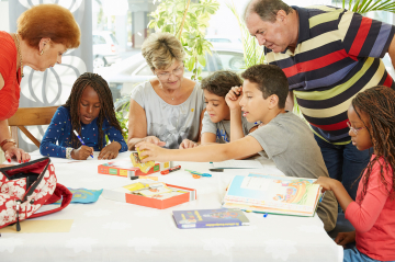 4 Kinder sitzen am Tisch mit Büchern und 3 Erwachsene helfen ihnen beim Lernen