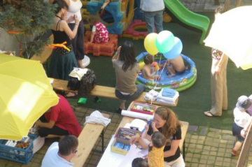 gelbe Sonnenschirme, Heurigentisch und Bänke, Erwachsenen und Kinder, die sprechen und spielen