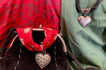 Bunte Trachtentücher mit Herzen-Anhängern, eine dunkle tracht-Ledertasche und im Hintergrund Blusen in rot und grün